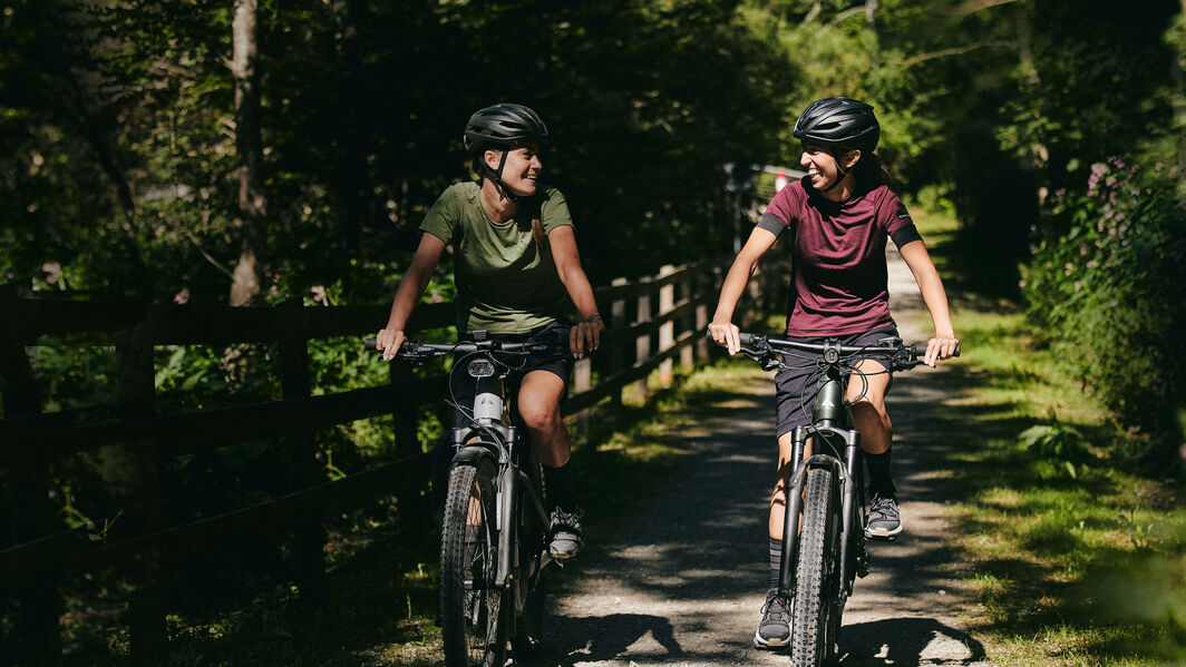 Two women riding Canyon e-touring bikes through a scenic, wooded countryside trail, enjoying a casual ride in nature.