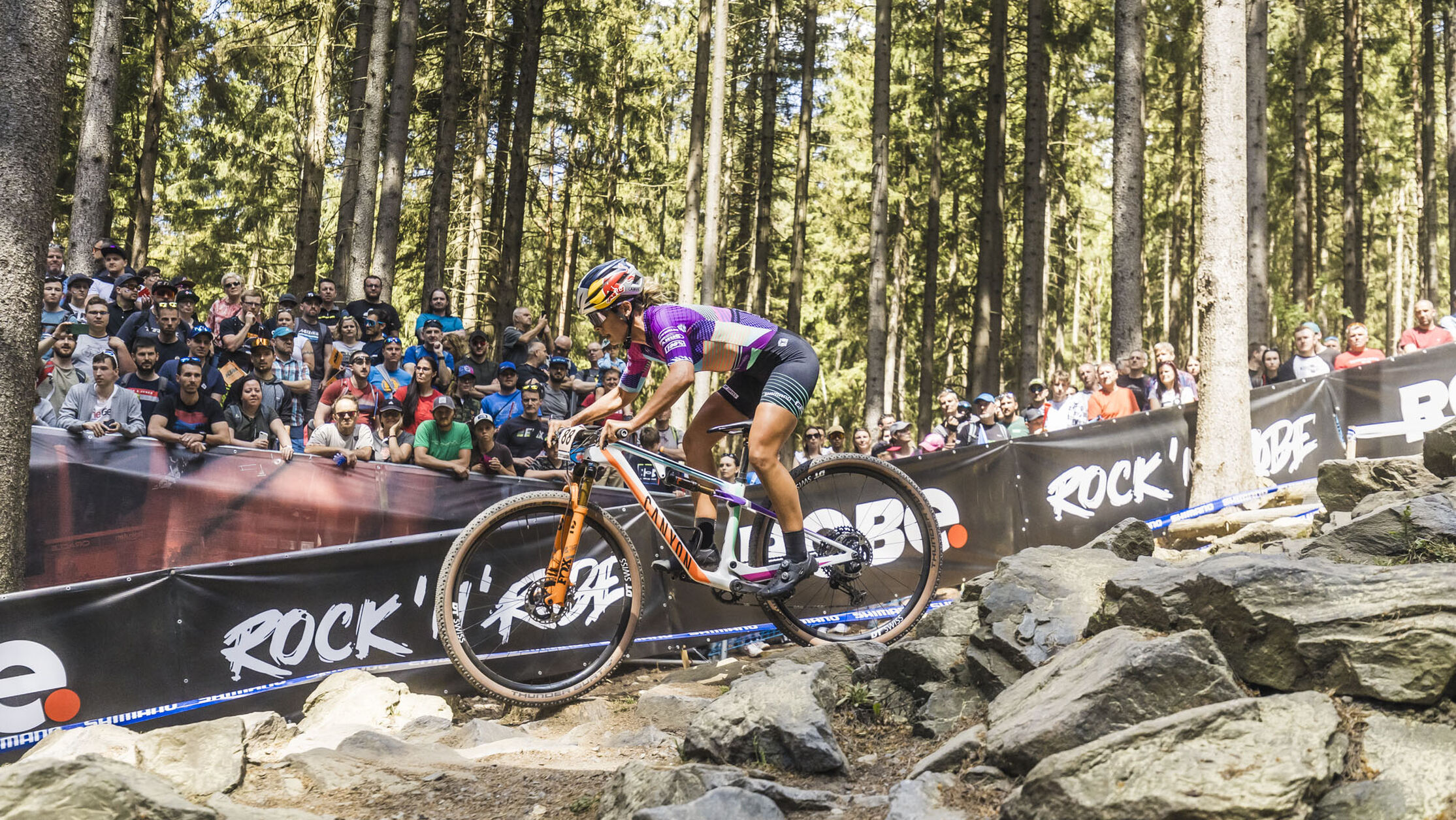 Canyon Lux cross-country mountain bike racing through rocky terrain with crowd watching in the background, equipped with 27.5 or 29-inch wheels.