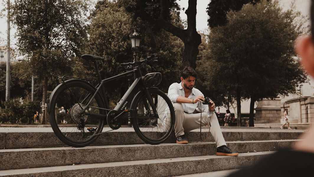 Explore the city in comfort with the perfect city bike — designed for smooth, everyday rides Man sitting on stone steps beside a Canyon bike, adjusting his camera in a park setting.